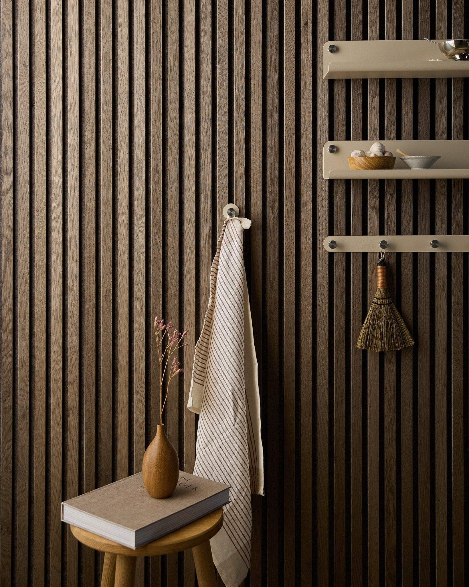 A towel hangs on a Norr Coat Hook by WoodUpp, mounted on an Akupanel acoustic wood slat wall beside beige shelves, with a broom, stool, vase, and book.